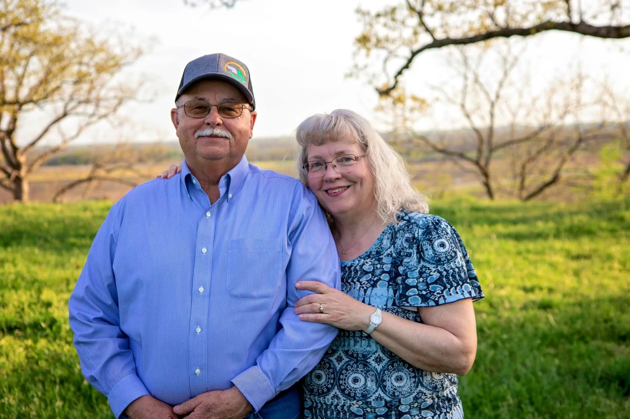 A happy couple stands together outdoors, with the man in a blue button-up shirt and cap, and the woman wearing a patterned blue top. They are surrounded by lush green grass and trees in the background, capturing a warm moment in nature.