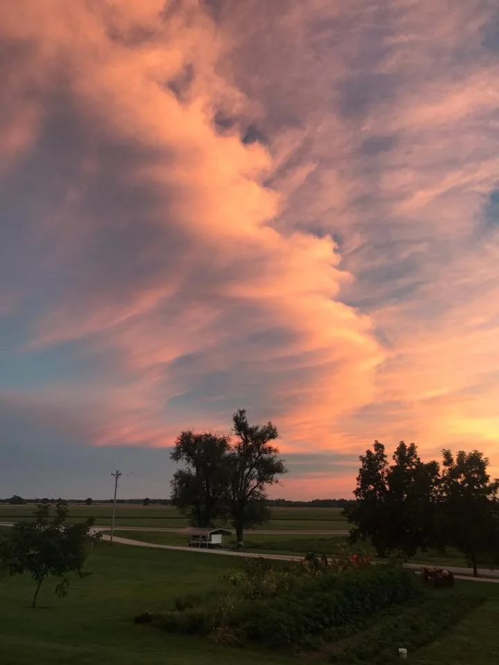 Vibrant sunset sky featuring colorful clouds in shades of pink and orange, with silhouettes of trees and a rural landscape in the foreground.