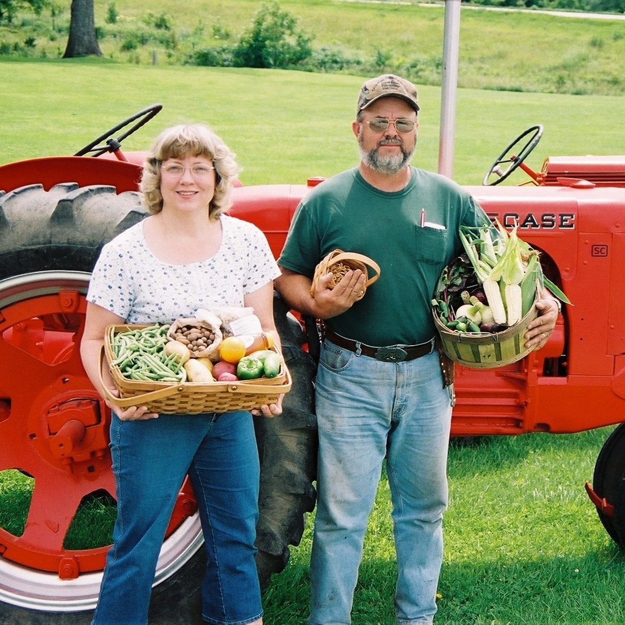 Two farmers standing in front of a red tractor, holding baskets of fresh vegetables and fruits. The woman on the left carries a basket filled with green beans, potatoes, and assorted fruits, while the man on the right holds a basket of corn and other produce. Lush green grass and a rural landscape are visible in the background, highlighting a farm setting.