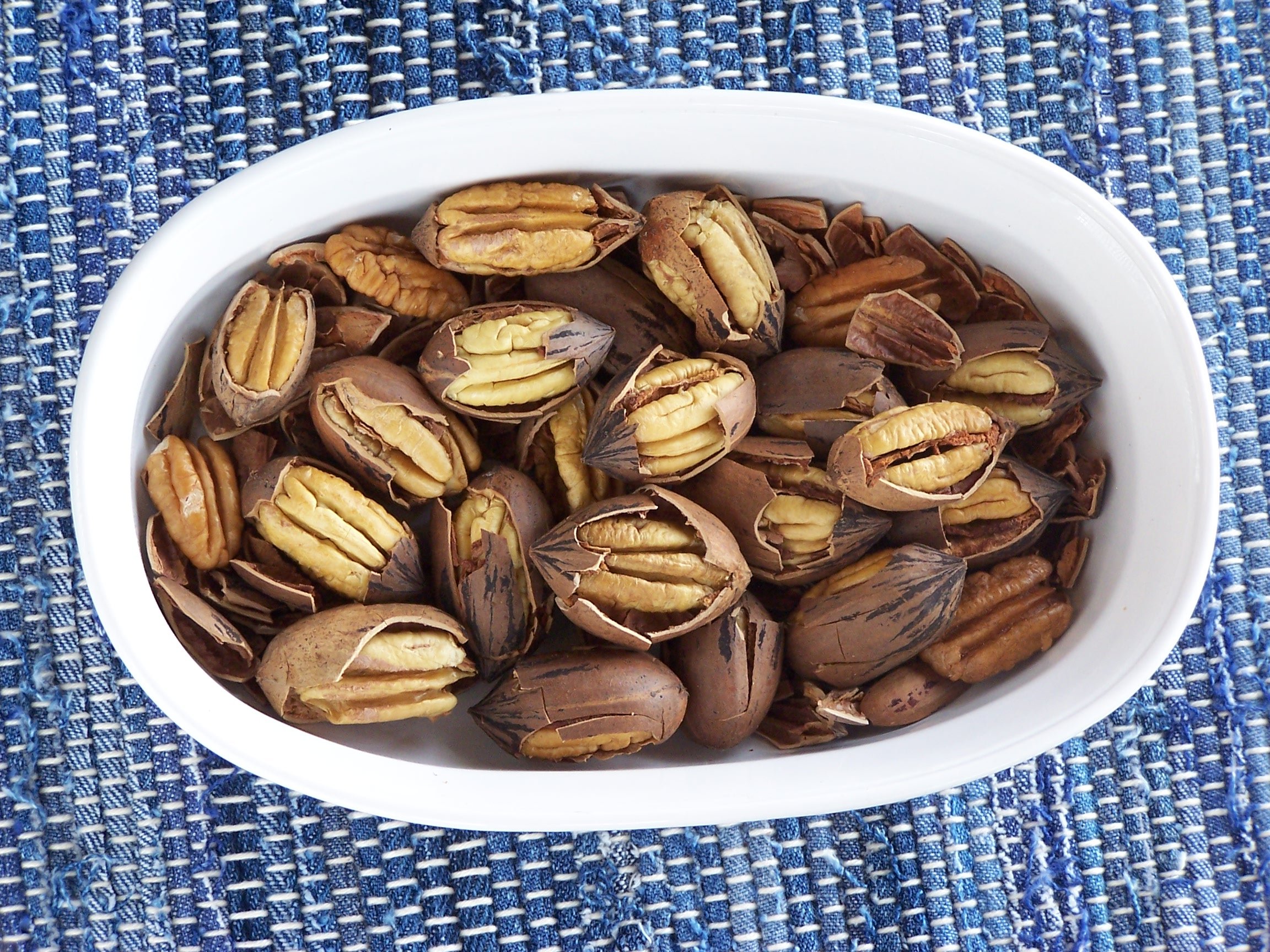Bowl of pecans in their shells, showcasing a mix of open and closed shells, on a blue woven textile background. Perfect for healthy snacking or baking.