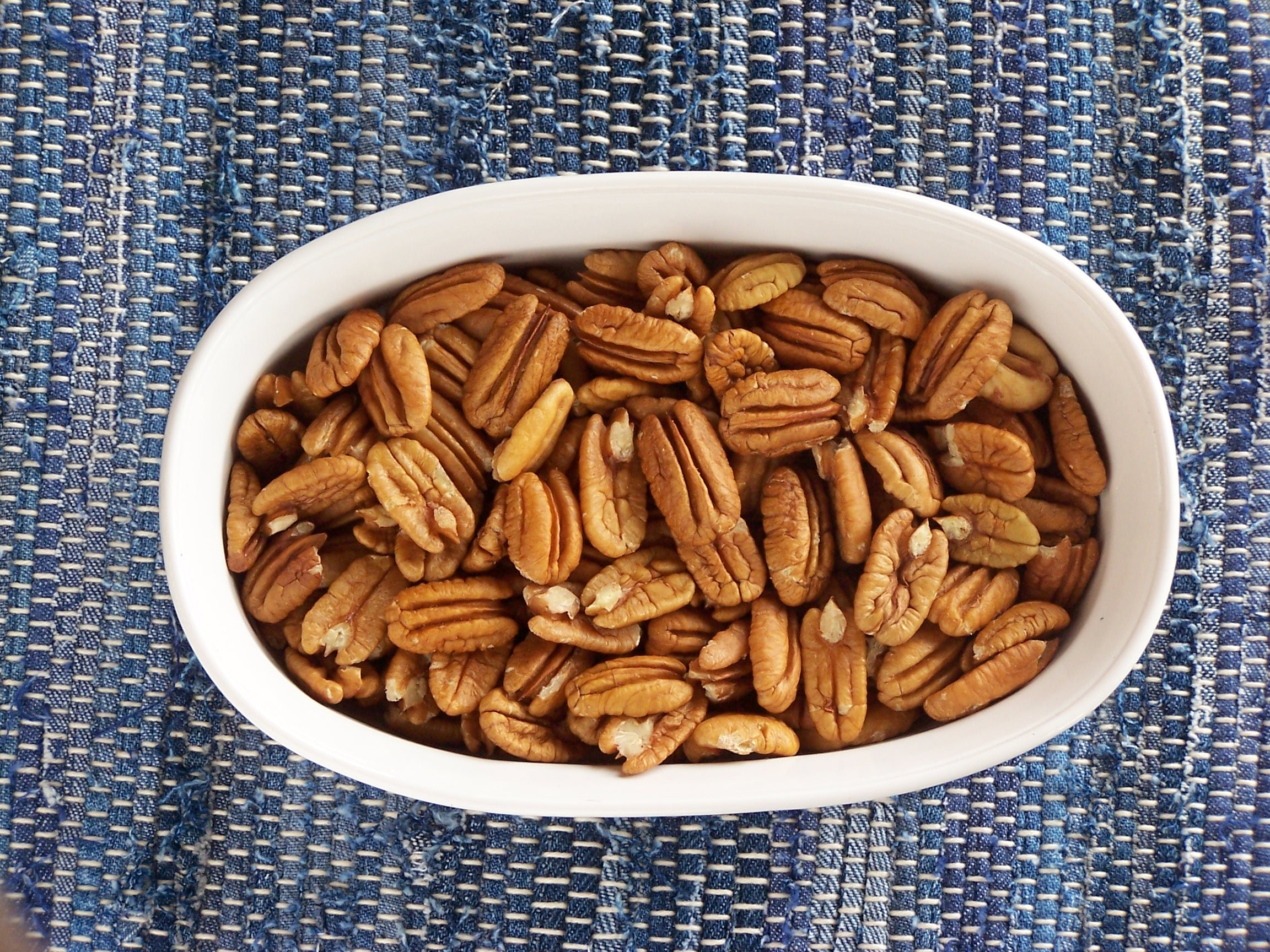 Bowl of roasted pecans on a textured blue woven tablecloth, showcasing their rich brown color and distinct shape, ideal for healthy snacking or cooking.