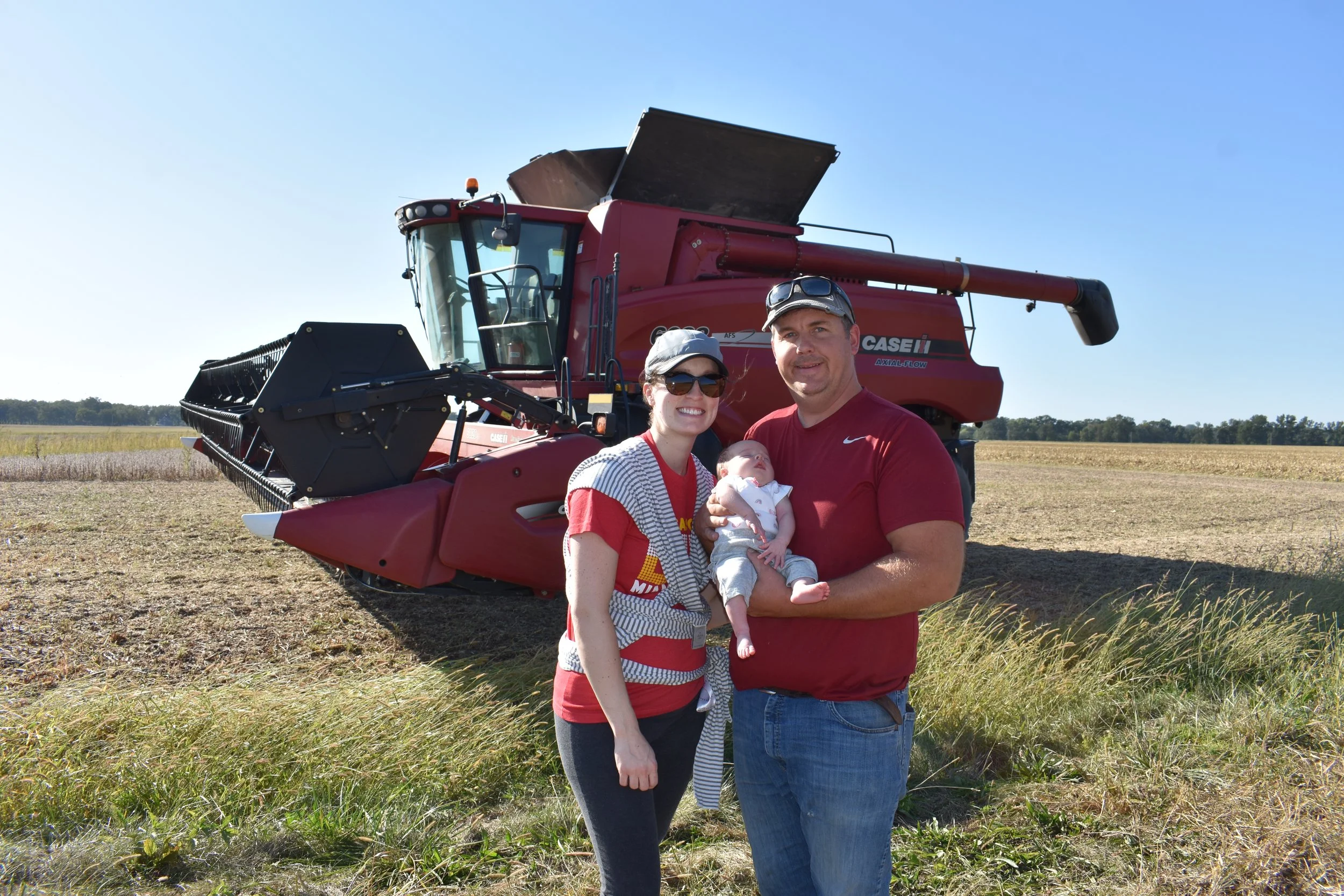Family standing in a field with a red Case IH combine harvester in the background, showcasing agricultural life and farming practices. The woman holds a baby while the man smiles, highlighting family involvement in farming. Clear blue sky and golden fields enhance the rural setting.