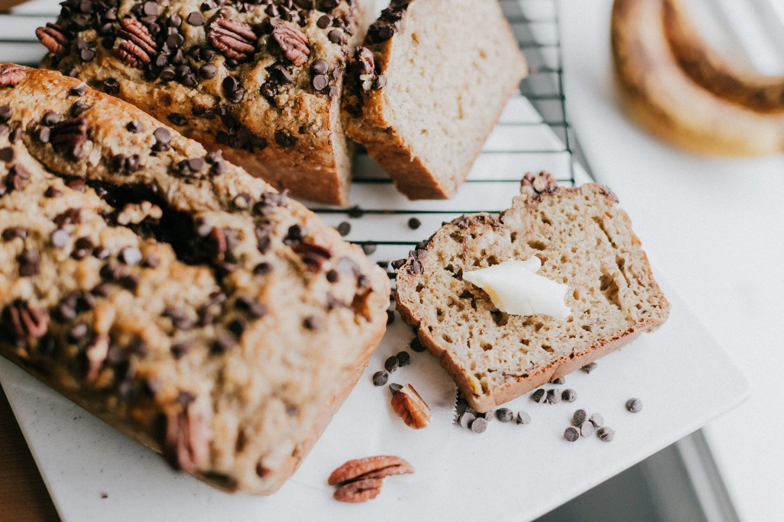 Sliced banana bread topped with chocolate chips and pecans, with a pat of butter on a white plate, surrounded by scattered chocolate chips and nuts.
